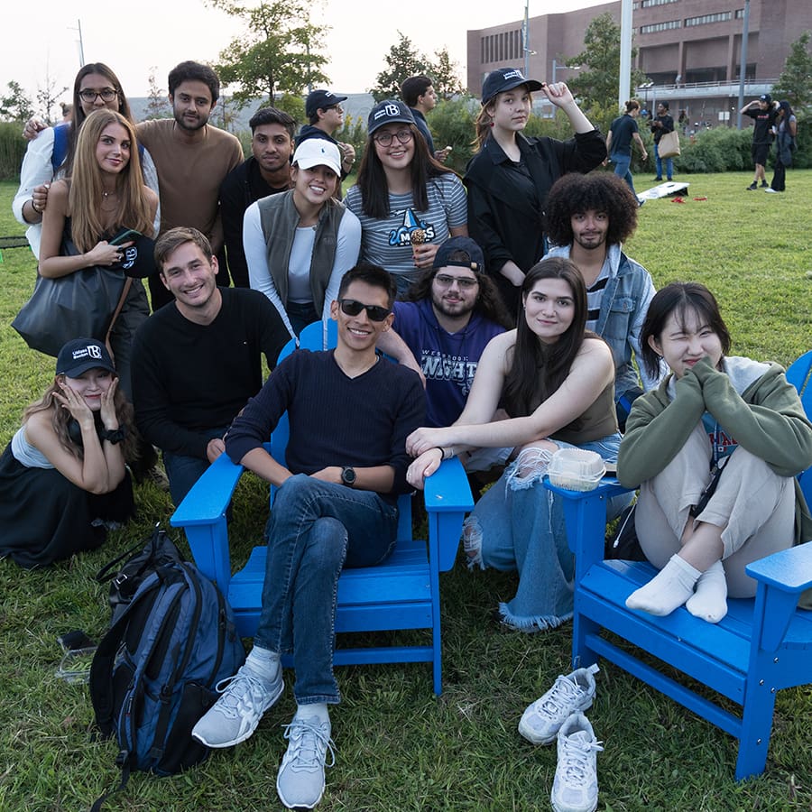 Group of students pose in blue chairs on campus center lawn.