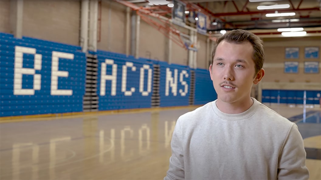 A young man stands in the UMass Boston gym where the bleachers say