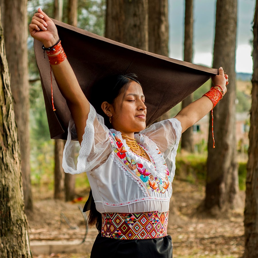 Young indigenous woman with traditional otavalo dress with her eyes closed holding a large scarf in her hands in the middle of a forest