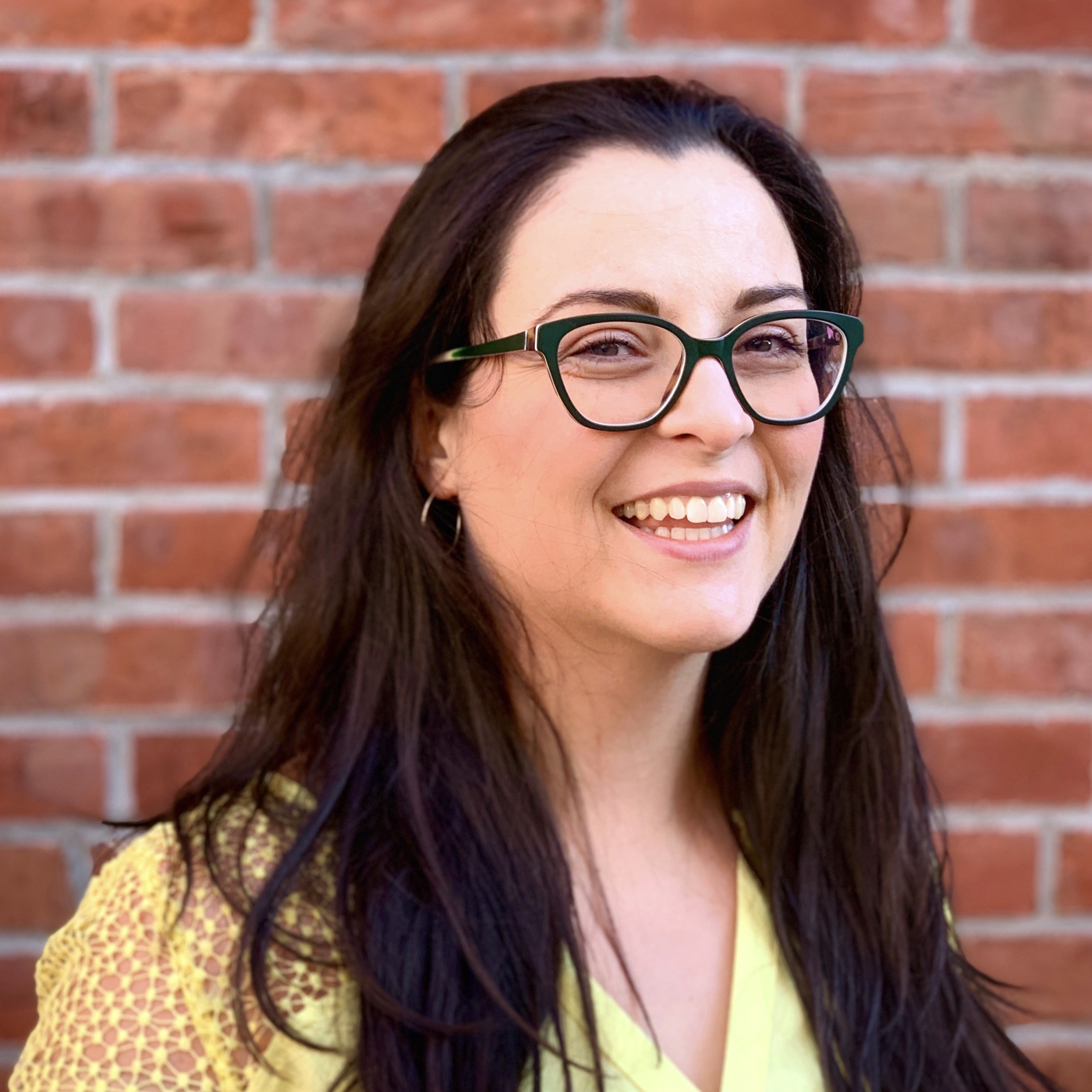 Woman with long brown hair, glasses, yellow shirt in front of brick wall